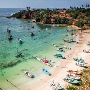 Vista aerea di numerose barche da pesca ormeggiate in una baia tropicale con acqua turchese cristallina e una spiaggia di sabbia.