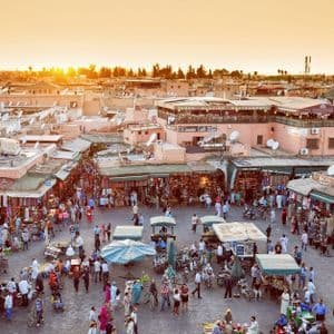 An elevated view of a bustling city marketplace with many people, stalls, and terracotta buildings at sunset.