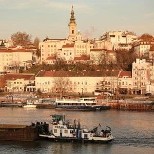 Un rimorchiatore manovra una chiatta su un fiume, con una città collinare e un campanile di chiesa prominente sullo sfondo.