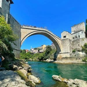 Prospettiva dal basso di un grande ponte ad arco in pietra che attraversa un fiume turchese, con persone riunite in cima sotto un cielo azzurro e limpido.