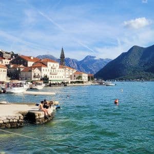 Due persone sedute su un molo di pietra che si affaccia su una baia con una città costiera e montagne sullo sfondo.