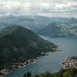 Una vista aerea di una baia sinuosa circondata da montagne verdi, con paesini lungo la riva e barche sull'acqua.