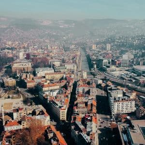 Una veduta aerea di una città fitta con edifici dai tetti rossi e un fiume, che si estende verso montagne sfumate all'orizzonte.