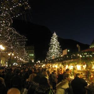 Una grande folla di persone cammina attraverso un mercatino di Natale all'aperto di notte, con bancarelle illuminate e alberi decorati.