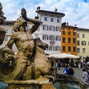 Una fontana in pietra con una statua di un tritone a cavallo di un cavalluccio marino in una piazza di città europea con edifici colorati.