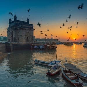 Pájaros sobrevuelan barcos en el agua, frente a un gran monumento de arco de piedra, durante un atardecer colorido.