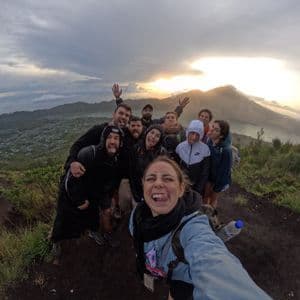 Un viaje en grupo de WeRoad se toma una selfie divertida con ojo de pez en la cima de una montaña al amanecer, con vistas a un volcán y un lago.