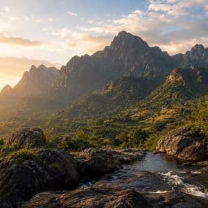 Un ruscello scende a cascata tra le rocce in una valle verde lussureggiante, con una catena montuosa sullo sfondo al tramonto.