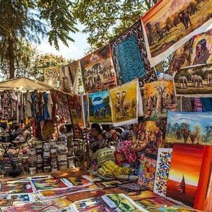 Una bancarella del mercato espone dipinti colorati di elefanti e paesaggi accanto a tessuti e altri prodotti artigianali sotto gli alberi.