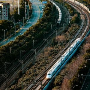 Veduta aerea di un treno proiettile bianco che viaggia su binari curvi, affiancato da un'autostrada tortuosa e altre linee ferroviarie.