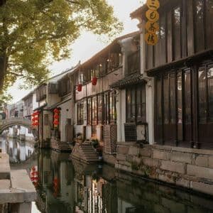 Edifici tradizionali con lanterne rosse costeggiano un canale calmo, con i loro riflessi che luccicano nell'acqua sotto un ponte in pietra ad arco.