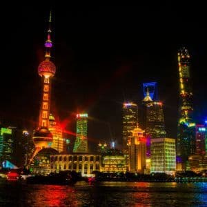Uno skyline moderno della città, con una torre distintiva e grattacieli illuminati da luci colorate che si riflettono sull'acqua di notte.