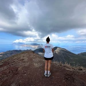 Un capogruppo WeRoad si trova su una cresta di montagna che domina un paesaggio vulcanico e il mare sotto un cielo nuvoloso.