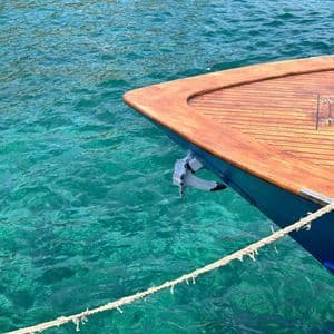 La prua di una barca con coperta in legno galleggia su acqua turchese trasparente, con l'ancora sollevata.