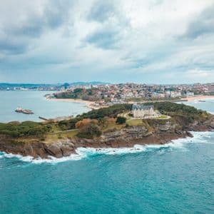 Una vista aérea de un palacio situado en una península arbolada que se adentra en el mar, con una ciudad costera y una playa en el fondo bajo un cielo nublado.