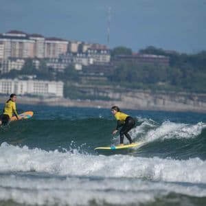 Dos personas de un viaje en grupo de WeRoad surfeando olas, con una en tabla de surf y una costa urbana de fondo.