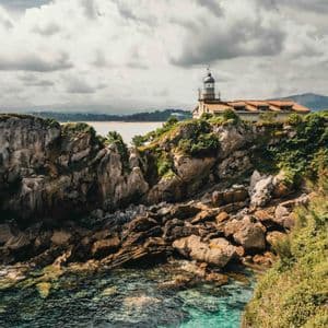 Un faro con techo de tejas se asienta en un acantilado rocoso y verde, con vistas a aguas claras y turquesas bajo un cielo nublado.
