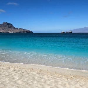 Ein Sandstrand mit leuchtend türkisfarbenem Wasser trifft auf ein tiefblaues Meer, mit felsigen Bergen und Schiffen in der Ferne.