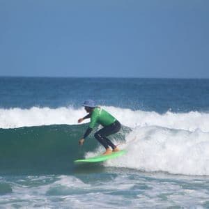 Un surfista con un traje de neopreno verde y negro y gorra azul surfea una ola en una tabla de surf verde brillante en el océano.