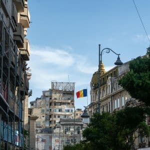 Eine rumänische Flagge hängt an einer Stadtstraße, umrahmt von alter, verzierter Architektur und einem weiteren Gebäude mit Gerüst.