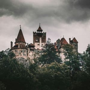 Eine mittelalterliche Burg mit roten Ziegeldächern und mehreren Türmen erhebt sich über einem dichten Wald unter einem dunklen, bewölkten Himmel.