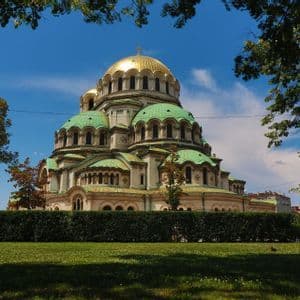 Eine große Kathedrale mit goldener Kuppel und grünen Dächern, von einem Park mit Hecke unter klarem blauem Himmel gesehen.