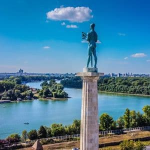 Eine Bronzestatue eines Kriegers auf einer hohen Säule blickt auf einen breiten Fluss, bewaldete Ufer und eine ferne Skyline unter blauem Himmel.