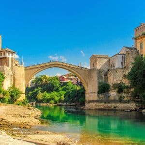 Eine gewölbte Steinbrücke überspannt einen türkisfarbenen Fluss und verbindet alte Steingebäude auf beiden Seiten unter einem klaren blauen Himmel.