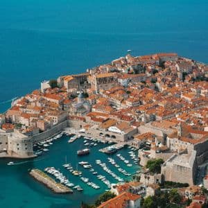 Luftaufnahme einer historischen, ummauerten Stadt mit orangefarbenen Ziegeldächern auf einer Halbinsel, die einen kleinen Hafen voller Boote im blauen Meer zeigt.