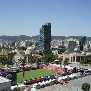 Eine Vogelperspektive einer Stadtlandschaft mit einer Fanzone auf einem Platz, die einen Fußballplatz, einen modernen Wolkenkratzer und ein Riesenrad zeigt.