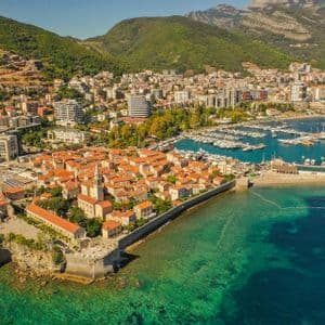 Eine Luftaufnahme einer ummauerten Küstenstadt auf einer Halbinsel, umgeben von türkisfarbenem Wasser, mit einem Yachthafen und Bergen in der Nähe.