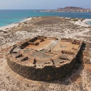 Vista aérea de las ruinas de un fuerte de piedra circular con cañones en una península arenosa, con océano turquesa y una ciudad distante.