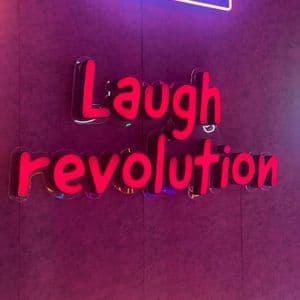 Un letrero rojo estilo neón dice 'Laugh revolution' en una pared morada texturizada, con una forma de neón azul encima.