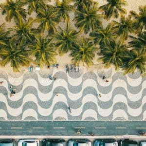 Vista aerea dall'alto di una spiaggia con palme, un marciapiede a mosaico ondulato, una strada e auto parcheggiate.