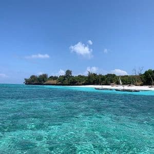 Acqua turchese cristallina in primo piano con barche a vela ormeggiate su una spiaggia di sabbia bianca, una costa alberata e un cielo blu.