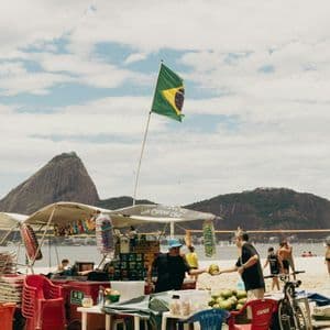 Un venditore su un carretto da spiaggia con una bandiera brasiliana serve una noce di cocco fresca a un cliente, con una montagna sullo sfondo.