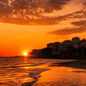 Un tramonto arancione si riflette sul mare e su una spiaggia di sabbia bagnata, con una città costiera stagliata su una scogliera sullo sfondo.