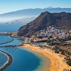 Una veduta aerea di una spiaggia affollata di sabbia dorata accanto a una località costiera, con montagne sullo sfondo sotto un cielo sereno.