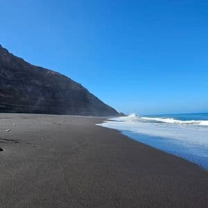 Weiße Wellen spülen an einen schwarzen Sandstrand neben einer steilen, felsigen Klippe unter klarem blauem Himmel.