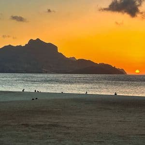 Menschen sitzen an einem Sandstrand und beobachten den Sonnenuntergang über dem Meer hinter einer schattenhaften Bergkette unter einem orangefarbenen Himmel.