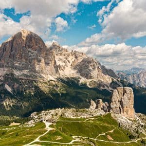 Una vista panoramica di una catena montuosa rocciosa, con cime prominenti, colline verdi con sentieri tortuosi e un cielo parzialmente nuvoloso.