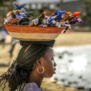 Una mujer de perfil con cabello trenzado equilibra una cesta tejida llena de artesanías coloridas sobre su cabeza junto al mar.