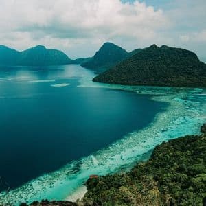 A high-angle view of lush, forested islands surrounded by turquoise and deep blue water, with a visible coral reef along the coastline.