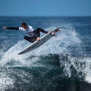 Un surfista con muta e maglia da competizione è in volo sulla sua tavola da surf, cavalcando la cresta di un'onda e creando spruzzi.