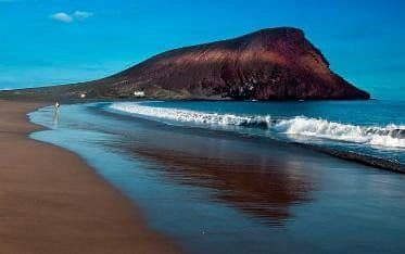 Una grande montagna rossastra domina una spiaggia di sabbia scura, con il suo riflesso proiettato sulla riva bagnata sotto un cielo azzurro chiaro.
