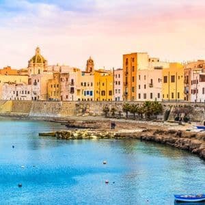 Edifici colorati di una città costiera si affacciano su una baia con acqua cristallina, con una piccola barca in primo piano durante l'alba.