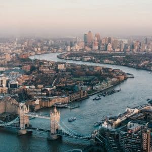An aerial view of Tower Bridge and the River Thames winding through a dense cityscape towards a modern skyline under a hazy sky.