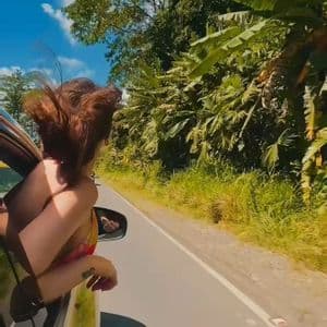 Una donna si sporge dal finestrino dell'auto con i capelli al vento, percorrendo una strada fiancheggiata da alberi verdi e lussureggianti.