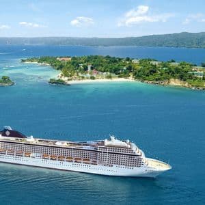 Una vista aerea di una grande nave da crociera bianca che naviga su un mare blu vicino a un'isola tropicale lussureggiante e verde.