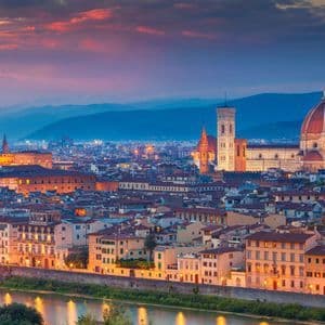 Una città illuminata con una grande cupola e un'alta torre lungo un fiume al tramonto, con montagne sullo sfondo.